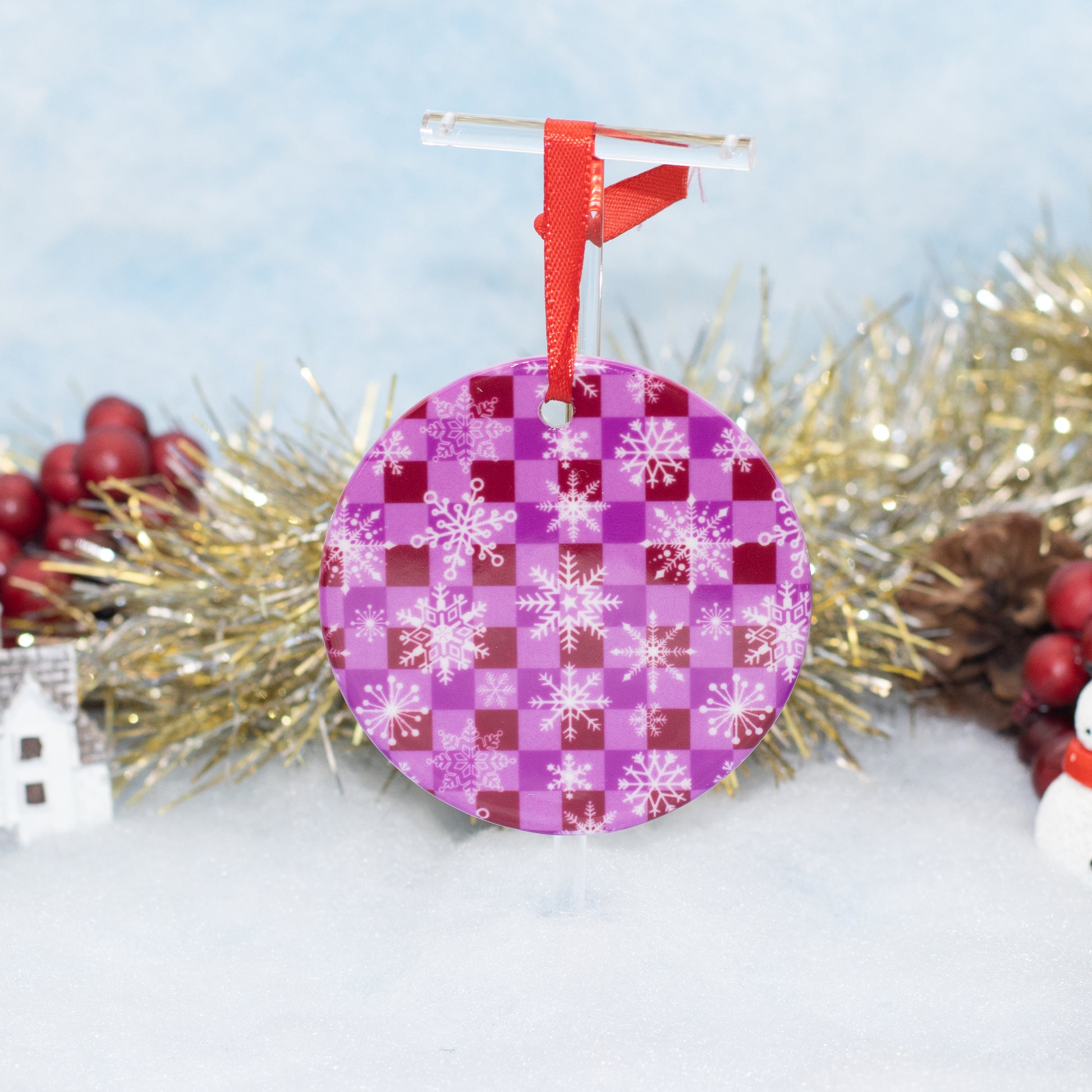 A round Christmas ornament with a purple and magenta checkerboard pattern and white snowflakes, hanging from a red ribbon and clear plastic hanger, styled with tinsel and red berries on a snowy background.