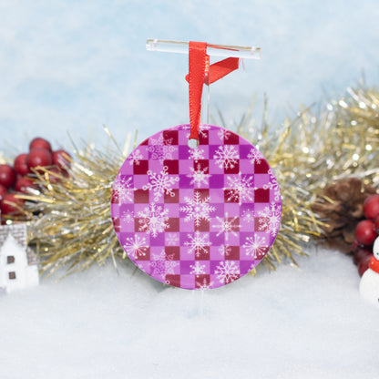 A round Christmas ornament with a purple and magenta checkerboard pattern and white snowflakes, hanging from a red ribbon and clear plastic hanger, styled with tinsel and red berries on a snowy background.