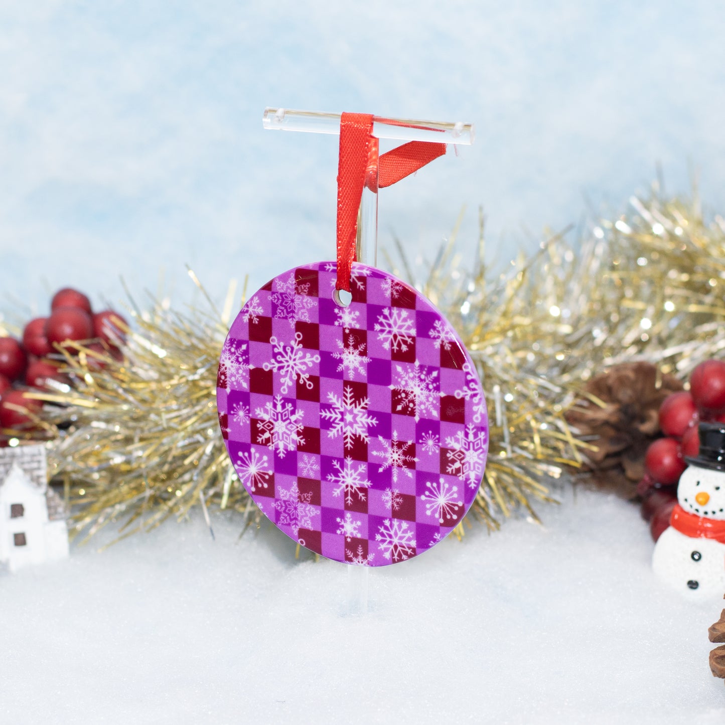 A round Christmas ornament with a purple and magenta checkerboard pattern and white snowflakes, hanging from a red ribbon on a clear stand, surrounded by festive tinsel, berries, and a small snowman decoration.