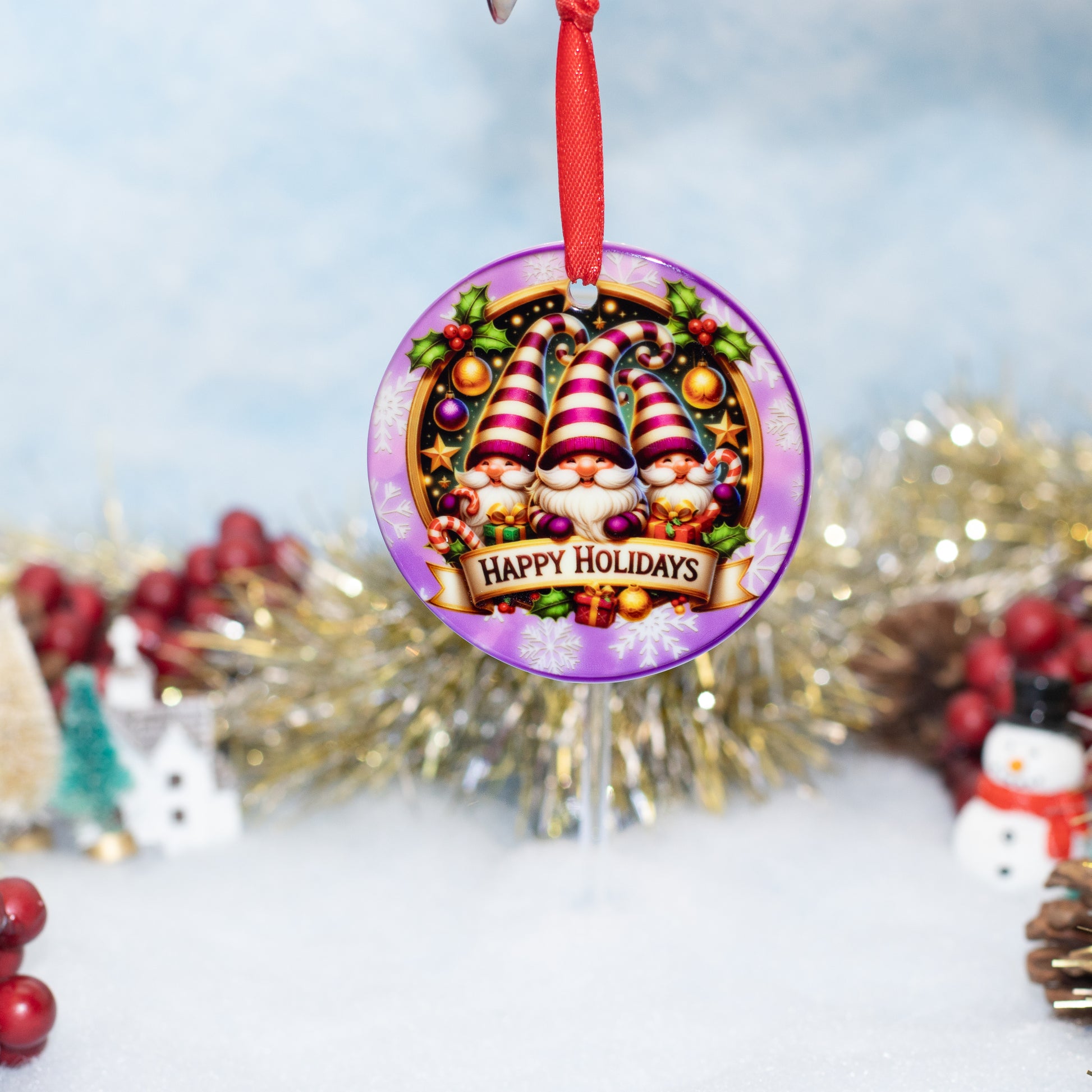 A round Christmas ornament featuring three festive gnomes with striped hats, surrounded by ornaments, berries, and a 'Happy Holidays' banner, hanging from a red ribbon against a softly blurred holiday background.
