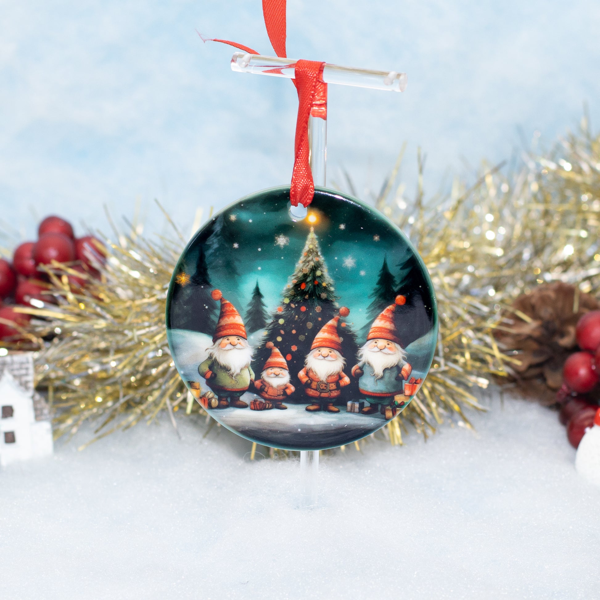 A round Christmas ornament featuring four gnome characters standing in front of a decorated Christmas tree, hanging from a red ribbon on a clear stand against a snowy outdoor-style holiday background with tinsel and berries.