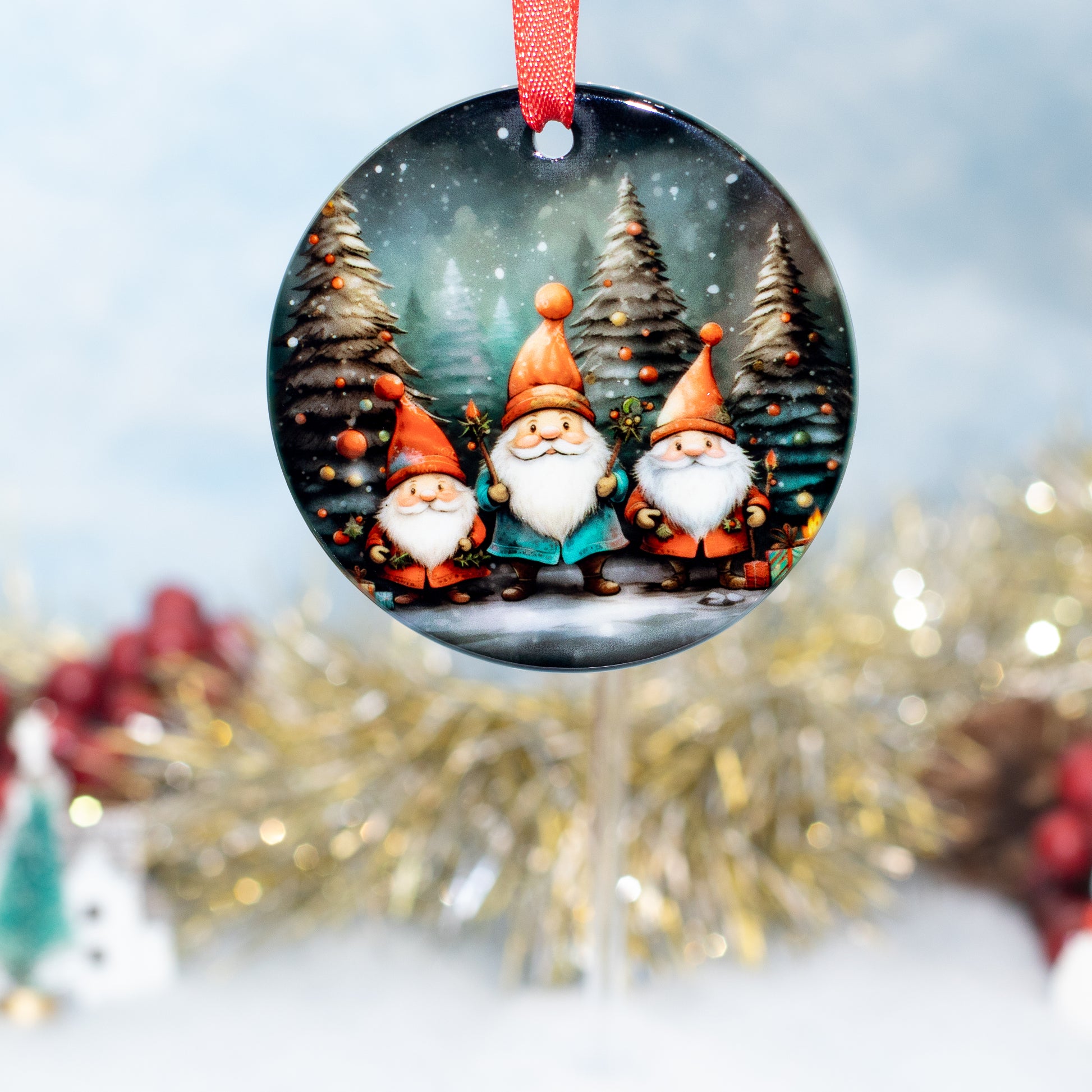 A round Christmas ornament featuring three festive gnomes in orange hats standing in a snowy pine tree landscape, hanging from a red ribbon against a softly blurred holiday background.