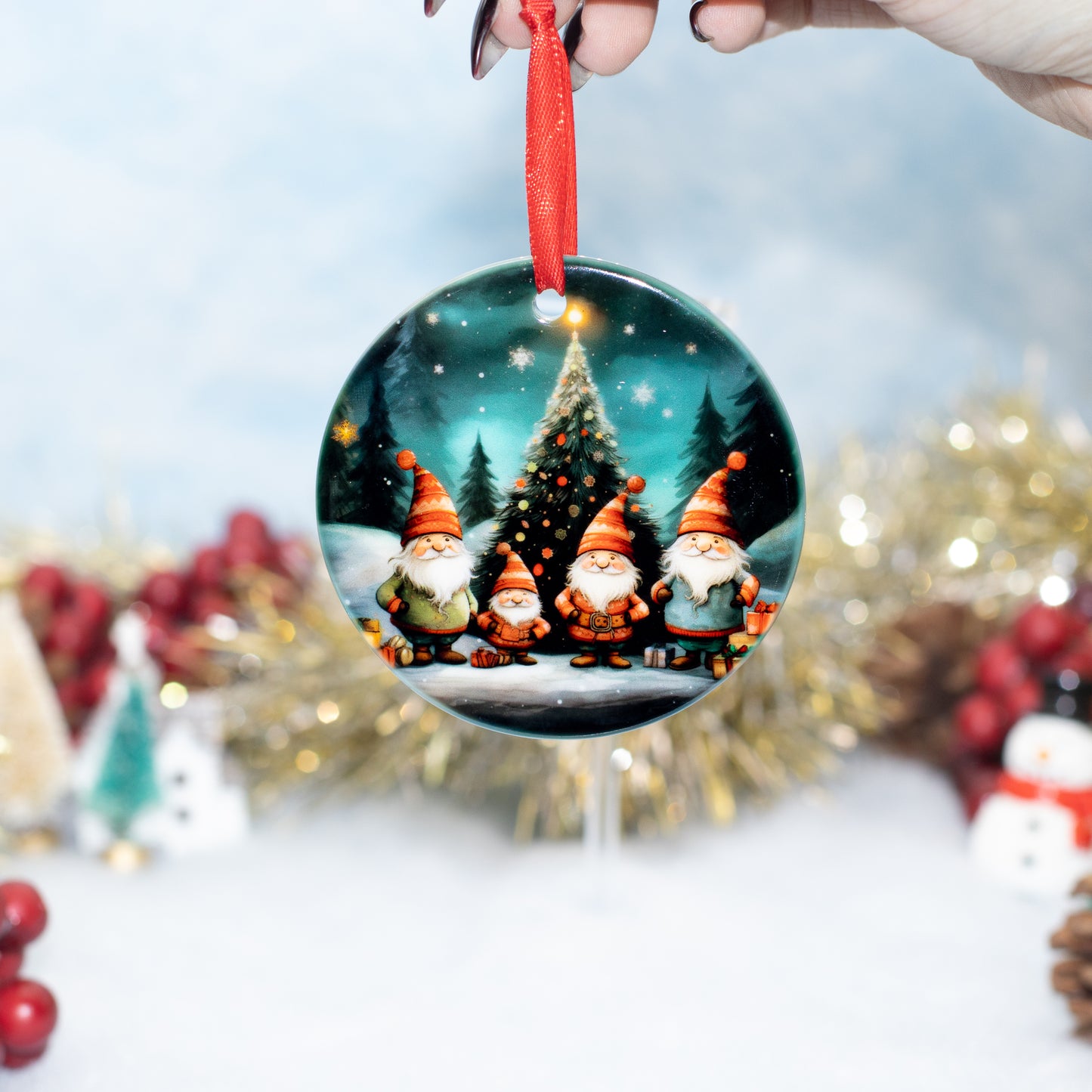 A round Christmas ornament hanging from a red ribbon held by a hand, featuring four festive gnomes standing in front of a glowing Christmas tree. The background is softly blurred with holiday decorations.
