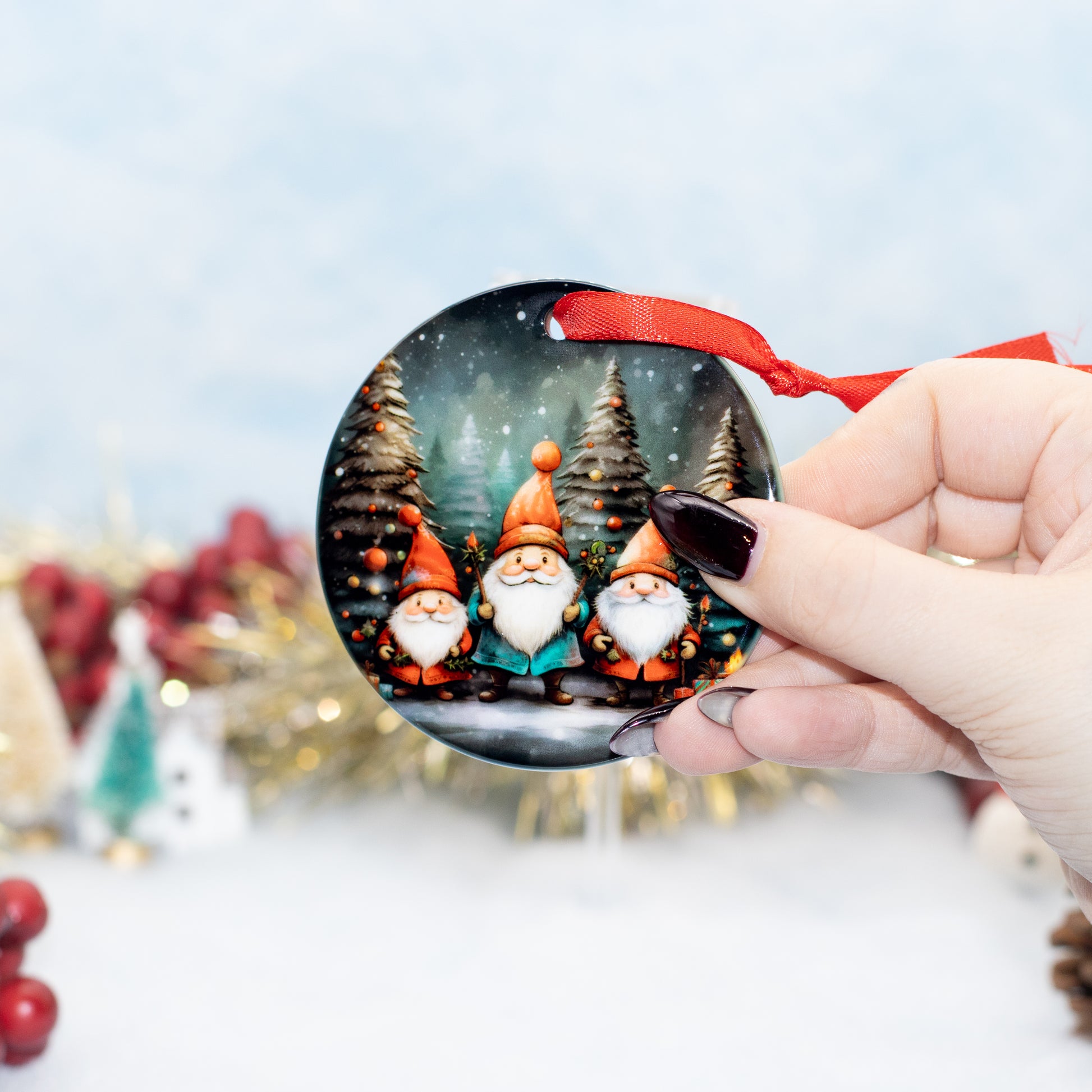 A hand holds a round Christmas ornament featuring three smiling gnomes in a snowy forest. The ornament has a red ribbon loop and is photographed against a softly blurred festive background with gold tinsel and red berries.