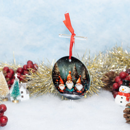 A round Christmas ornament featuring three festive gnomes in front of pine trees, hanging from a clear stand with a red ribbon. The ornament is styled on a snowy surface with gold tinsel, red berries, a miniature house, and a snowman figurine against a soft blue background.