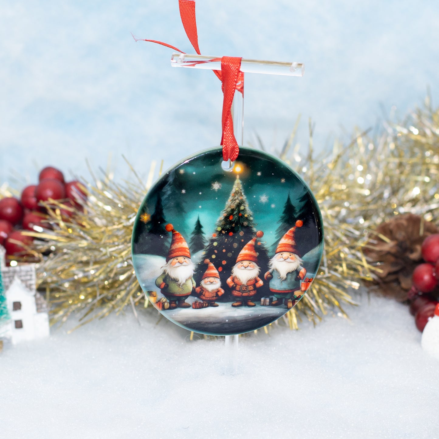 A round Christmas ornament featuring three festive gnomes standing in front of a glowing Christmas tree, hanging from a clear stand with a red ribbon. The ornament is styled on a white surface with gold tinsel, red berries, and a soft blue background.