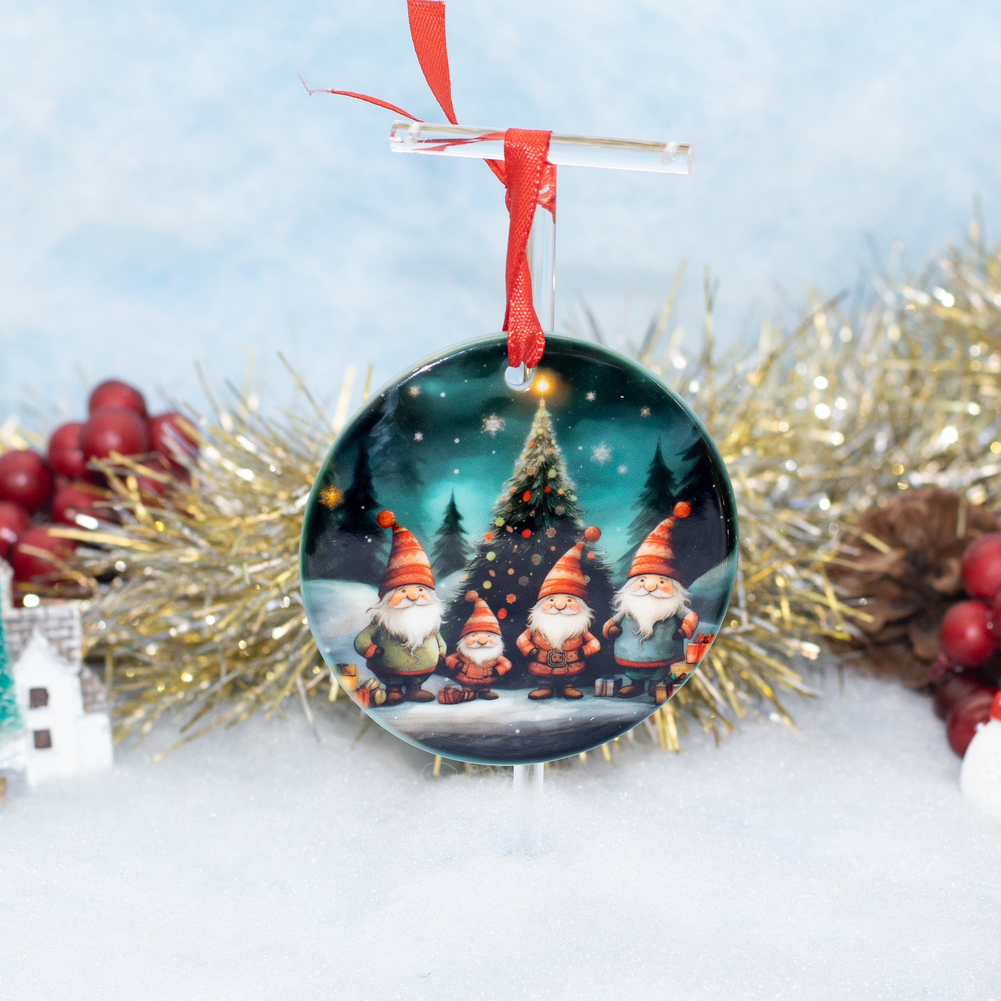 A round Christmas ornament featuring three festive gnomes standing in front of a glowing Christmas tree, hanging from a clear stand with a red ribbon. The ornament is styled on a white surface with gold tinsel, red berries, and a soft blue background.