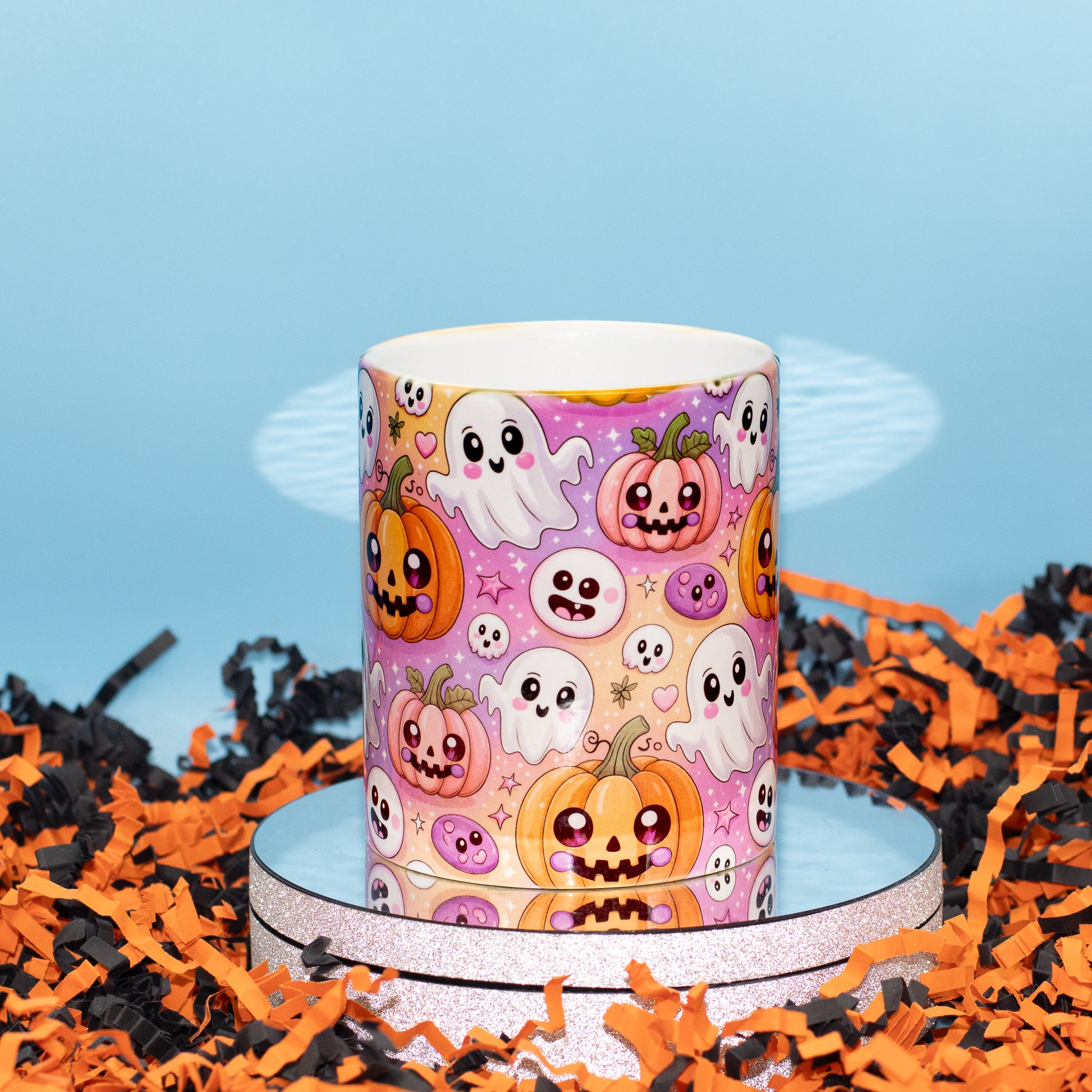 A ceramic mug with a colourful Halloween pattern featuring cartoon pumpkins, ghosts, and skulls on a pink background, displayed on a round silver tray surrounded by orange and black shredded paper against a light blue backdrop.
