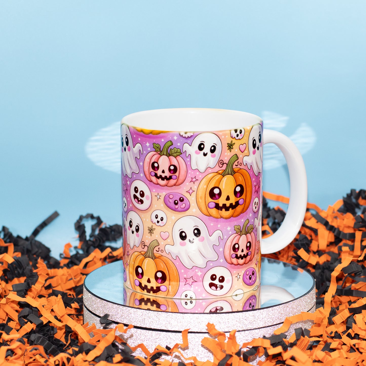 A white ceramic mug with a colorful Halloween print featuring pumpkins, ghosts, and skulls, displayed on a mirrored stand surrounded by orange and black shredded paper against a light blue background.