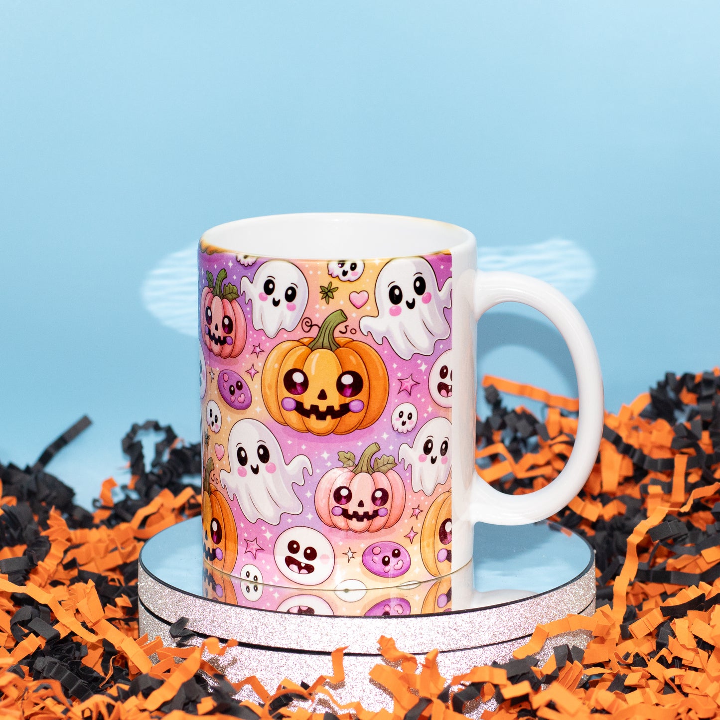 A white ceramic mug with a colorful Halloween pattern featuring cute pumpkins, ghosts, and skulls, displayed on a round silver stand amidst orange and black shredded paper against a light blue background.