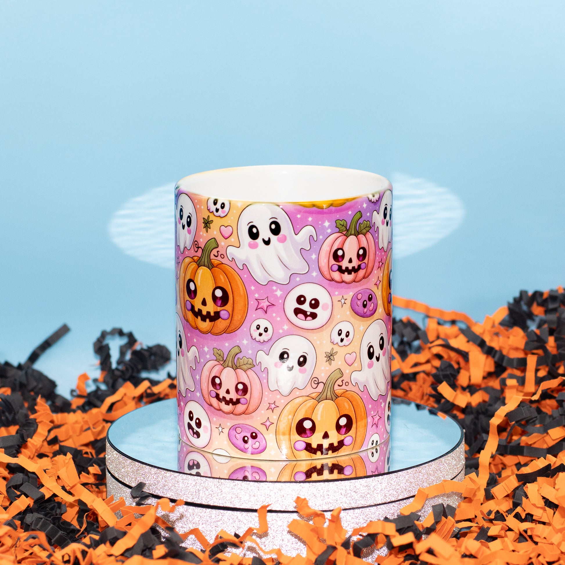 A Ceramic mug with a colourful Halloween pattern featuring cartoon pumpkins, ghosts, and skulls on a pink background, displayed on a round pedestal with orange and black shredded paper around it.