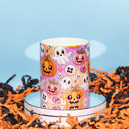 A Ceramic mug with a colourful Halloween pattern featuring cartoon pumpkins, ghosts, and skulls on a pink background, displayed on a round pedestal with orange and black shredded paper around it.