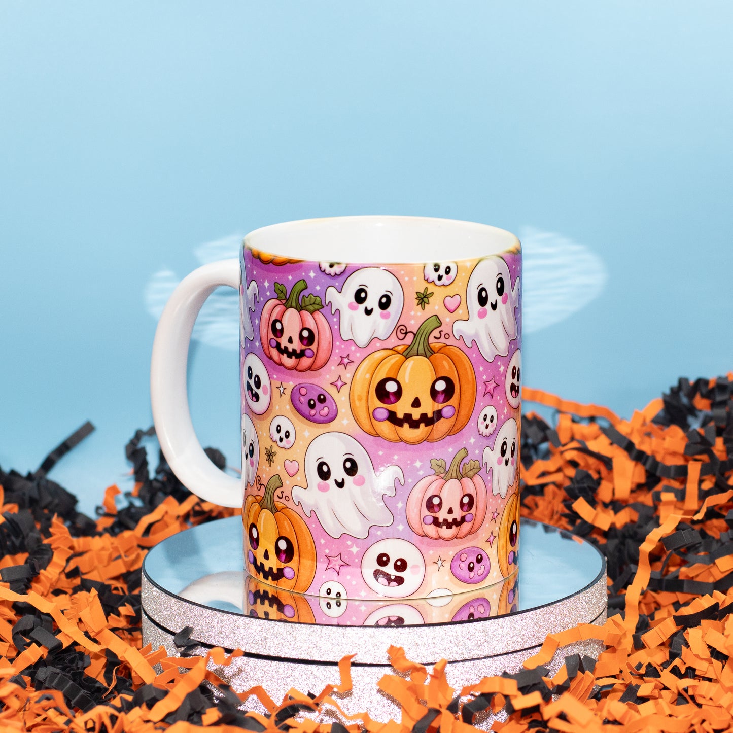 A white ceramic mug with a colorful Halloween print featuring pumpkins, ghosts, and skulls on a pink and purple background, displayed on a round silver riser surrounded by orange and black shredded paper against a light blue backdrop.