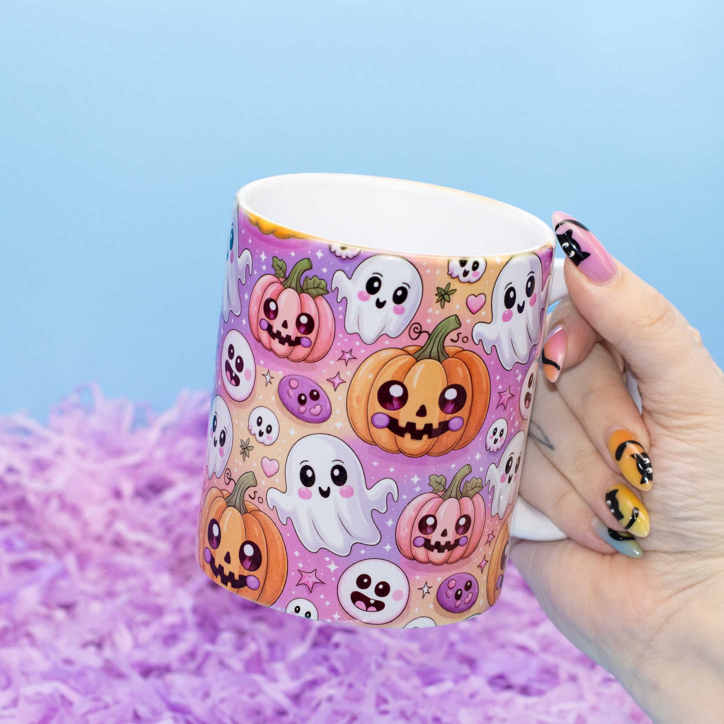 white ceramic mug decorated with a cute, colorful pattern of smiling pumpkins, ghosts, and skulls on a purple background, set against a light blue backdrop with purple shredded paper.