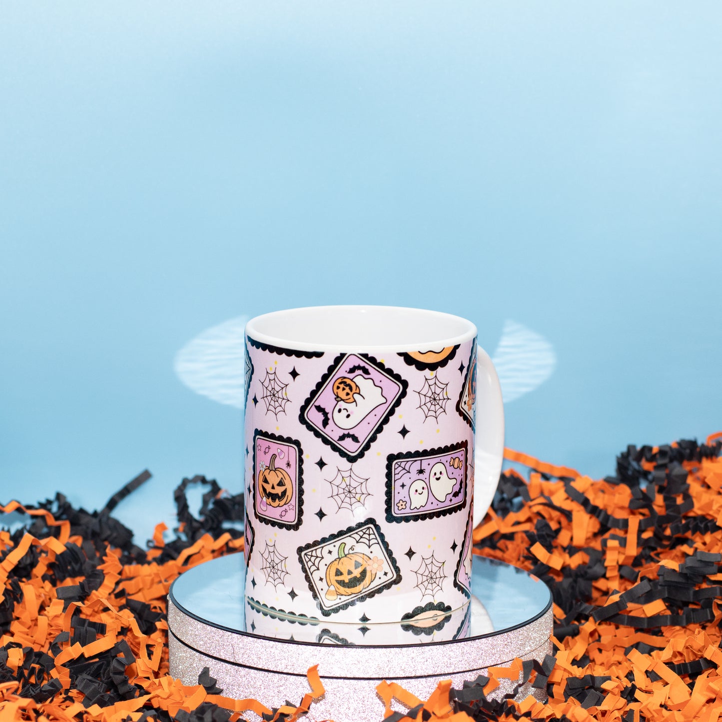 A white ceramic mug with a Halloween-themed pattern of jack-o'-lanterns, ghosts, and spiderwebs, displayed on a mirrored stand surrounded by orange and black shredded paper against a light blue background.