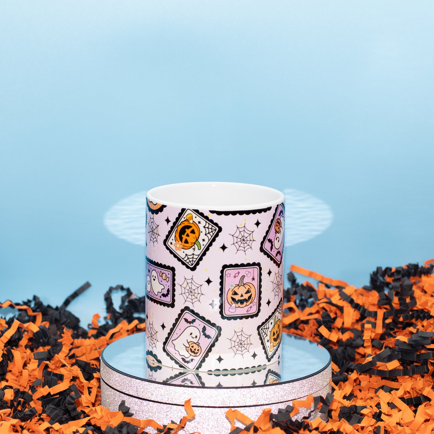 A white ceramic mug with a Halloween-themed pattern of jack-o'-lanterns, ghosts, and spiderwebs, displayed on a silver reflective stand surrounded by orange and black shredded paper against a light blue background.