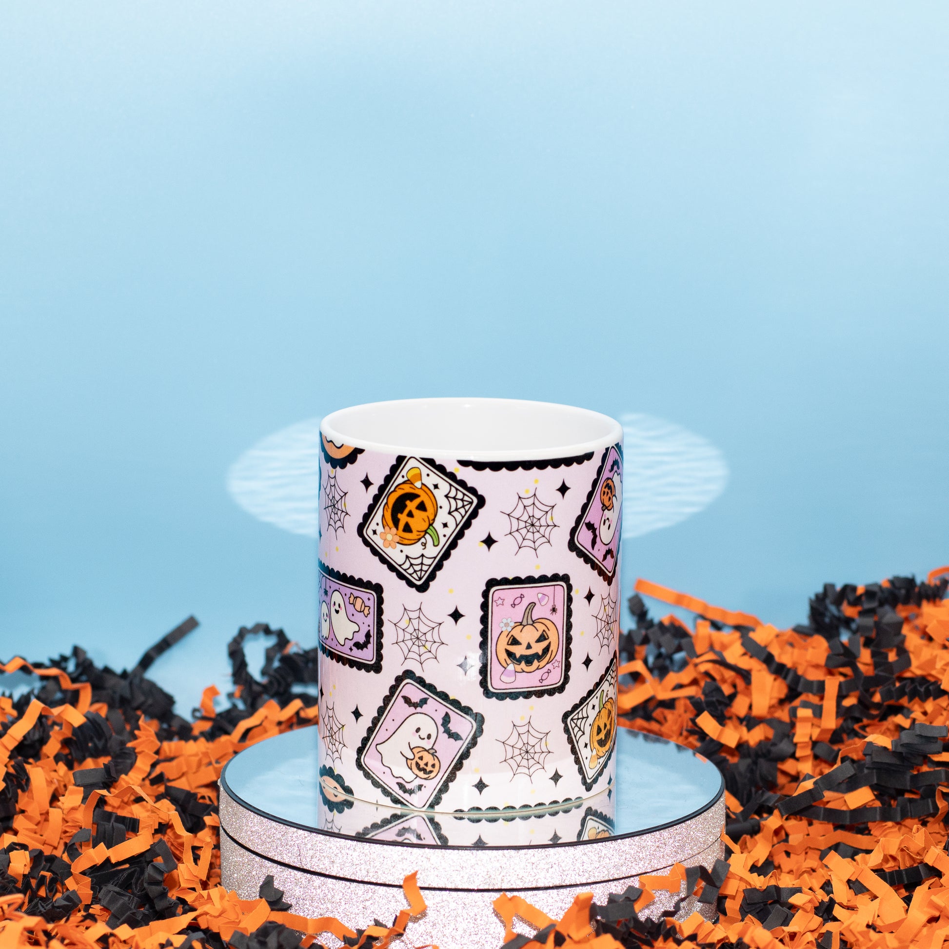 A white ceramic mug with a Halloween-themed pattern of jack-o'-lanterns, ghosts, and spiderwebs, displayed on a silver reflective stand surrounded by orange and black shredded paper against a light blue background.