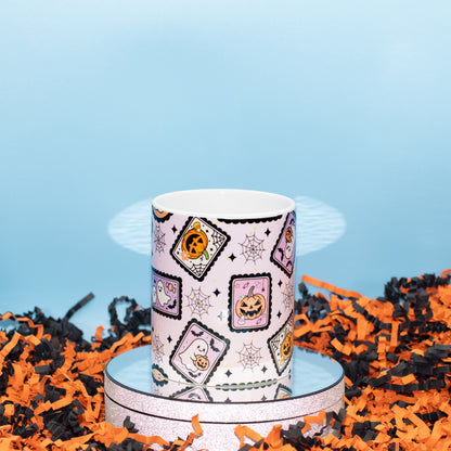 A white ceramic mug with a Halloween-themed pattern of jack-o'-lanterns, ghosts, and spiderwebs, displayed on a silver reflective stand surrounded by orange and black shredded paper against a light blue background.