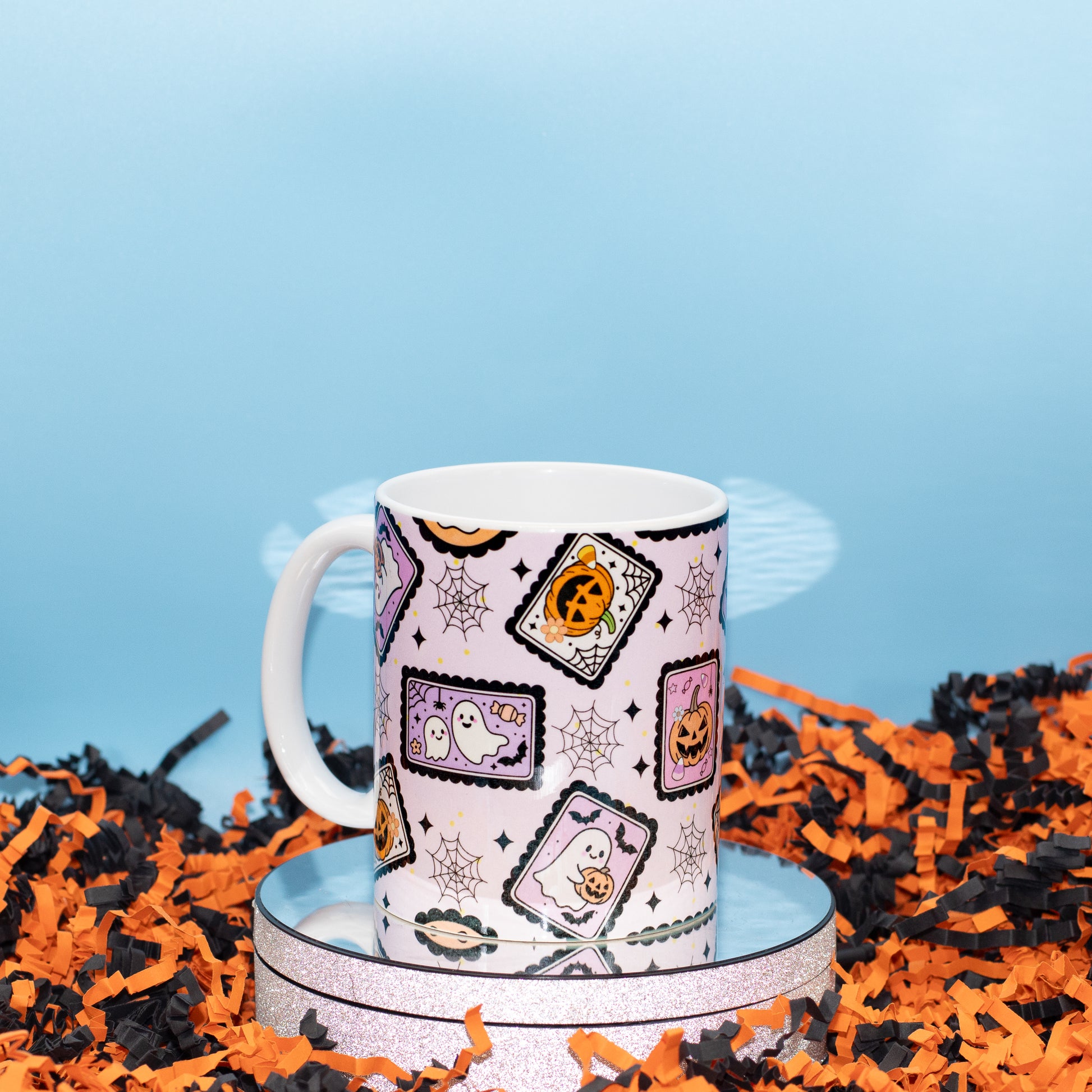 A white ceramic mug with a Halloween-themed pattern of ghostly postcards, spiderwebs, and jack-o'-lanterns, displayed on a round silver tray surrounded by orange and black shredded paper against a light blue background.