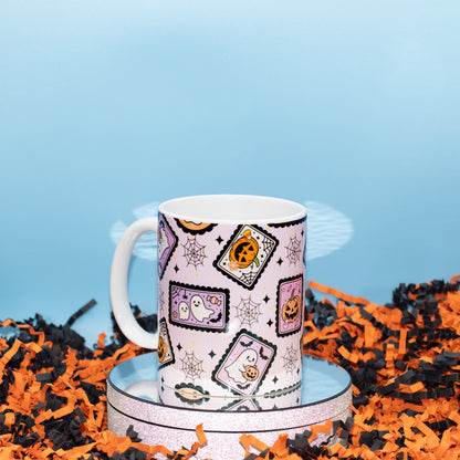 A white ceramic mug with a Halloween-themed pattern of ghostly postcards, spiderwebs, and jack-o'-lanterns, displayed on a round silver tray surrounded by orange and black shredded paper against a light blue background.
