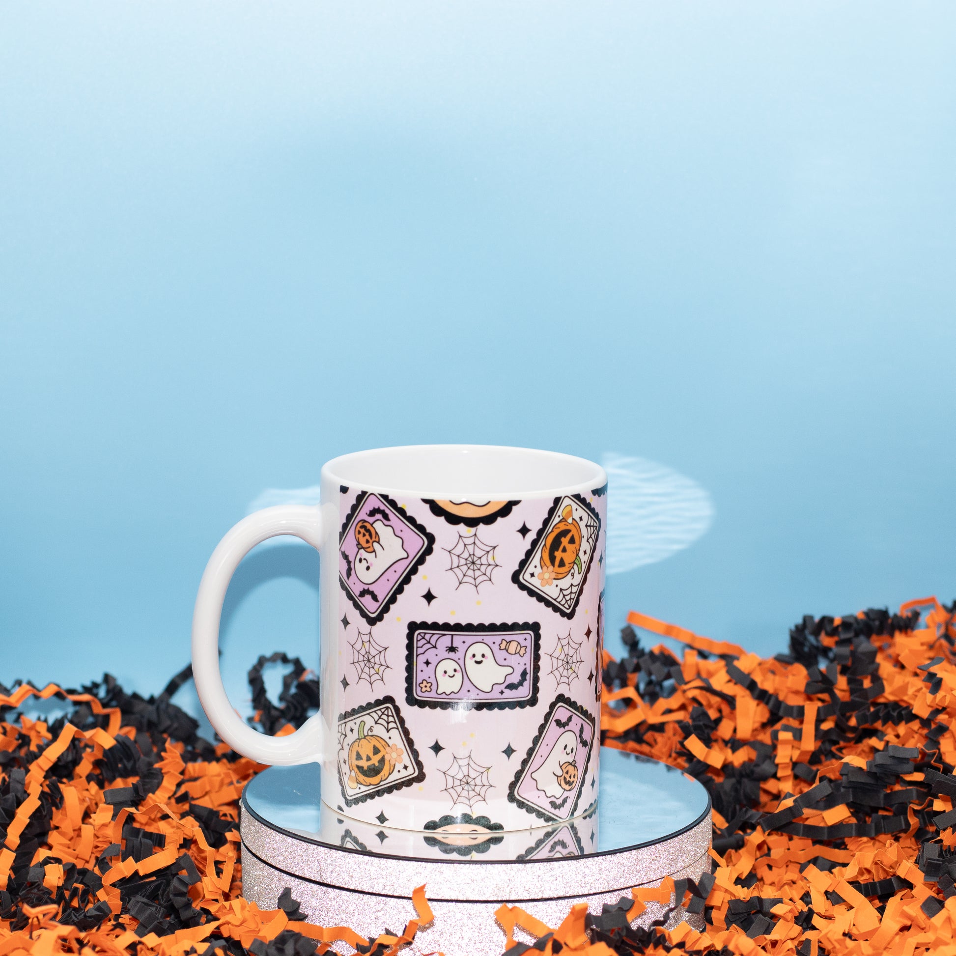 A white ceramic mug with a Halloween-themed pattern of tombstones, ghosts, and spiderwebs, displayed on a reflective silver tray surrounded by orange and black shredded paper against a light blue background.