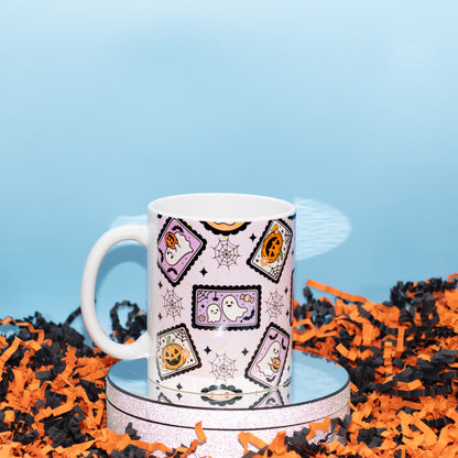 A white ceramic mug with a Halloween-themed pattern of candy corn, ghosts, and jack-o'-lanterns on a light pink background, displayed on a round silver tray surrounded by orange and black shredded paper against a light blue background.