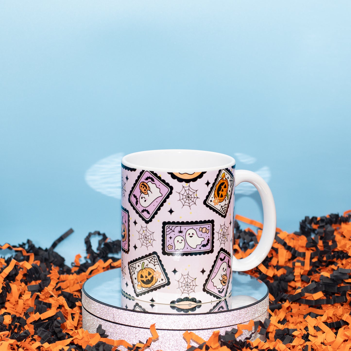 A white ceramic mug with a Halloween-themed pattern of candy corn, ghosts, and jack-o'-lanterns on a light pink background,