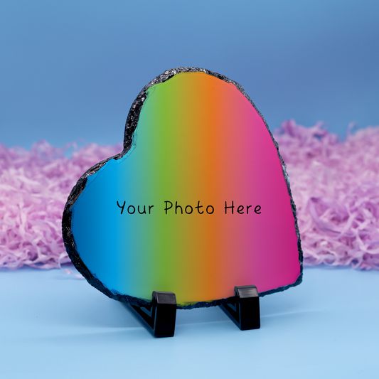 A heart-shaped photo slate stone with a vibrant rainbow gradient, displayed on a small black stand against a blue background with pink shredded paper.