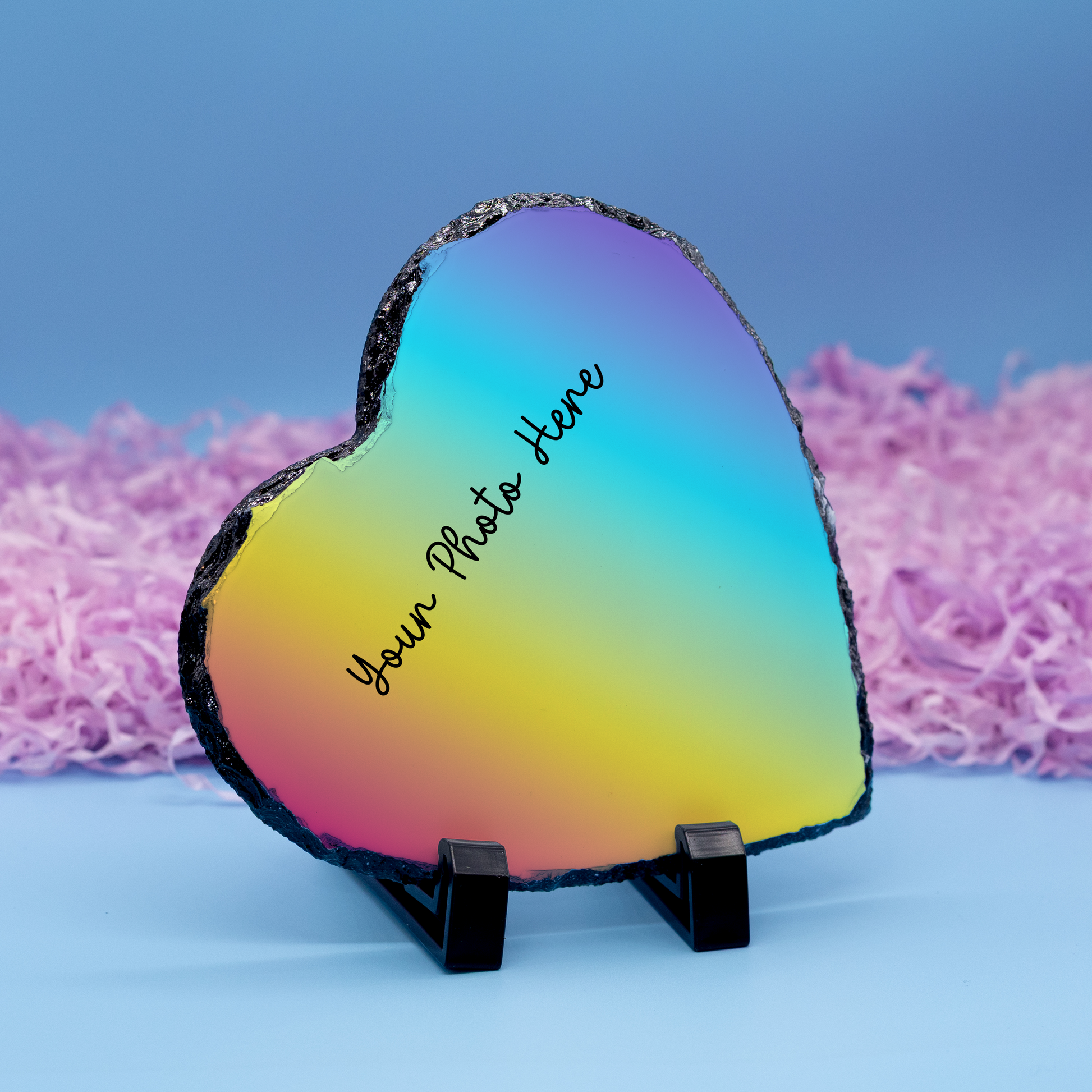 A heart-shaped polished stone plaque with a vibrant rainbow gradient, displayed on a small black stand against a blue background with pink shredded paper. The stone features a black textured rim and a customizable photo placeholder printed in black script.