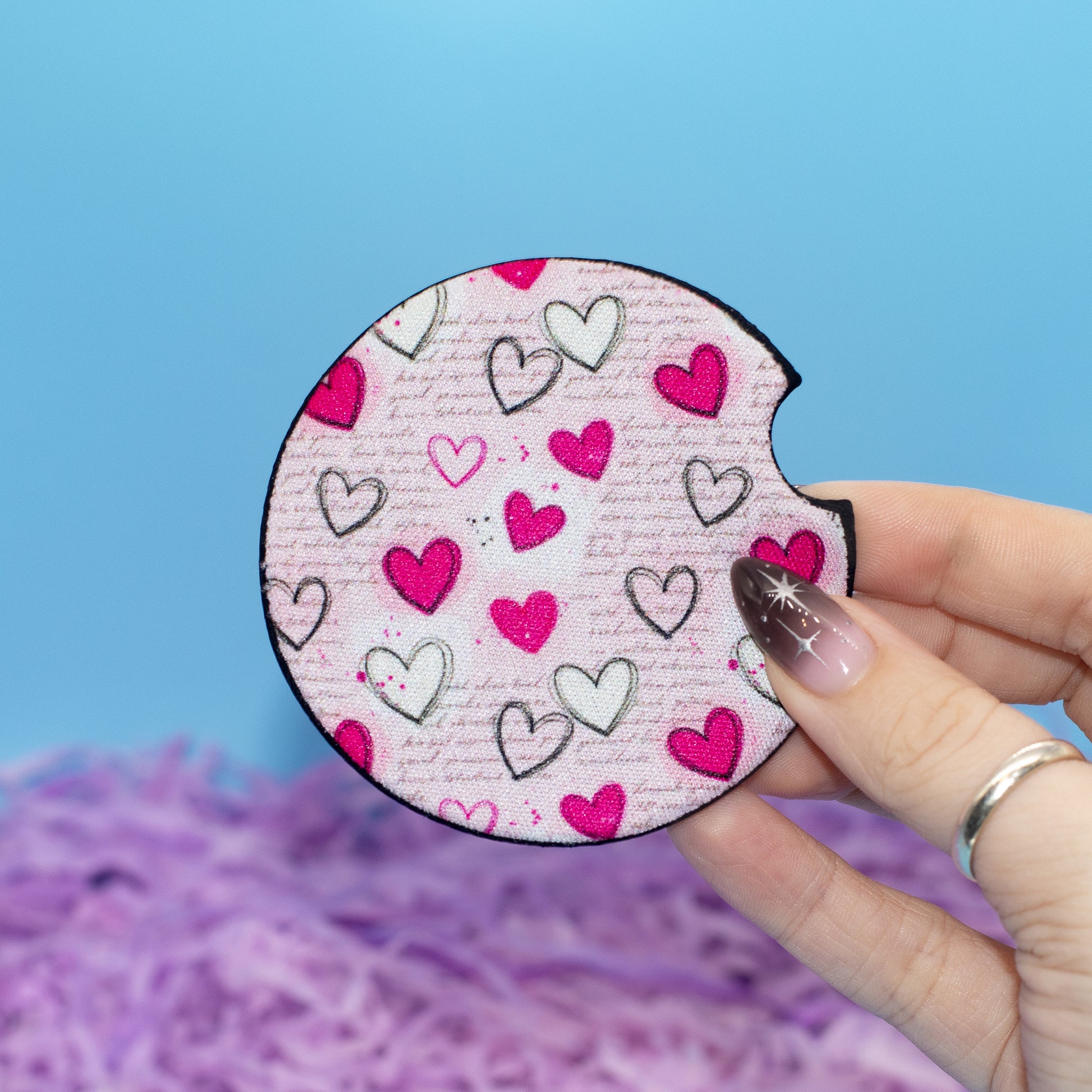 A hand holds a car coaster featuring a pink and white heart pattern, set against a bright blue background with purple shredded paper at the bottom.