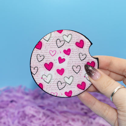 A hand holds a car coaster featuring a pink and white heart pattern, set against a bright blue background with purple shredded paper at the bottom.