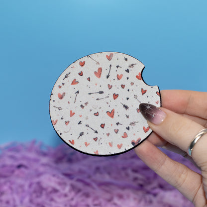 A hand holds a car coaster  featuring a white background with a pattern of small red hearts, black arrows, and feathers. The background consists of a solid blue wall and purple fluffy decorative paper.