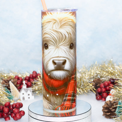 A tall stainless steel  tumbler with a straw, printed with a detailed portrait of a highland cow wearing a red plaid scarf, displayed on a silver pedestal with holiday decorations in the background.