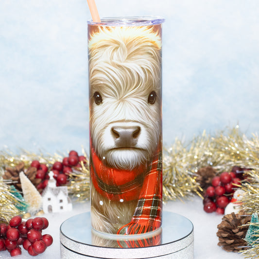 A tall stainless steel  tumbler with a straw, printed with a detailed portrait of a highland cow wearing a red plaid scarf, displayed on a silver pedestal with holiday decorations in the background.