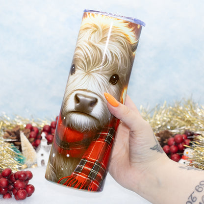 A hand holds a stainless steel tumbler featuring a printed illustration of a Scottish Highland cow wearing a red plaid scarf. The background is decorated with gold tinsel, red berries, and a light blue textured backdrop.