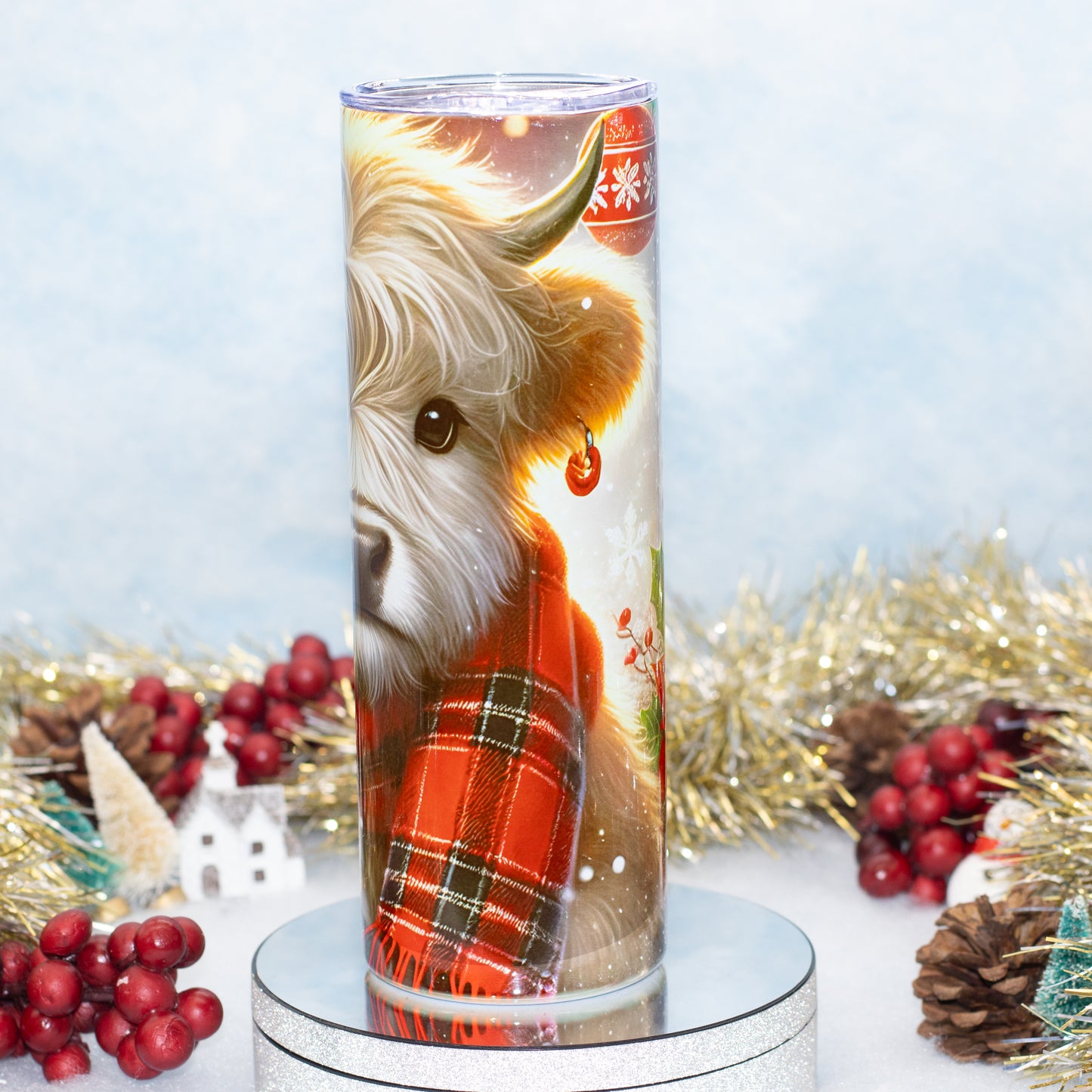 A tall cylindrical tumbler with a clear lid, featuring a printed illustration of a fluffy Highland cow wearing a red plaid scarf, displayed on a round silver pedestal with holiday decorations in the background.