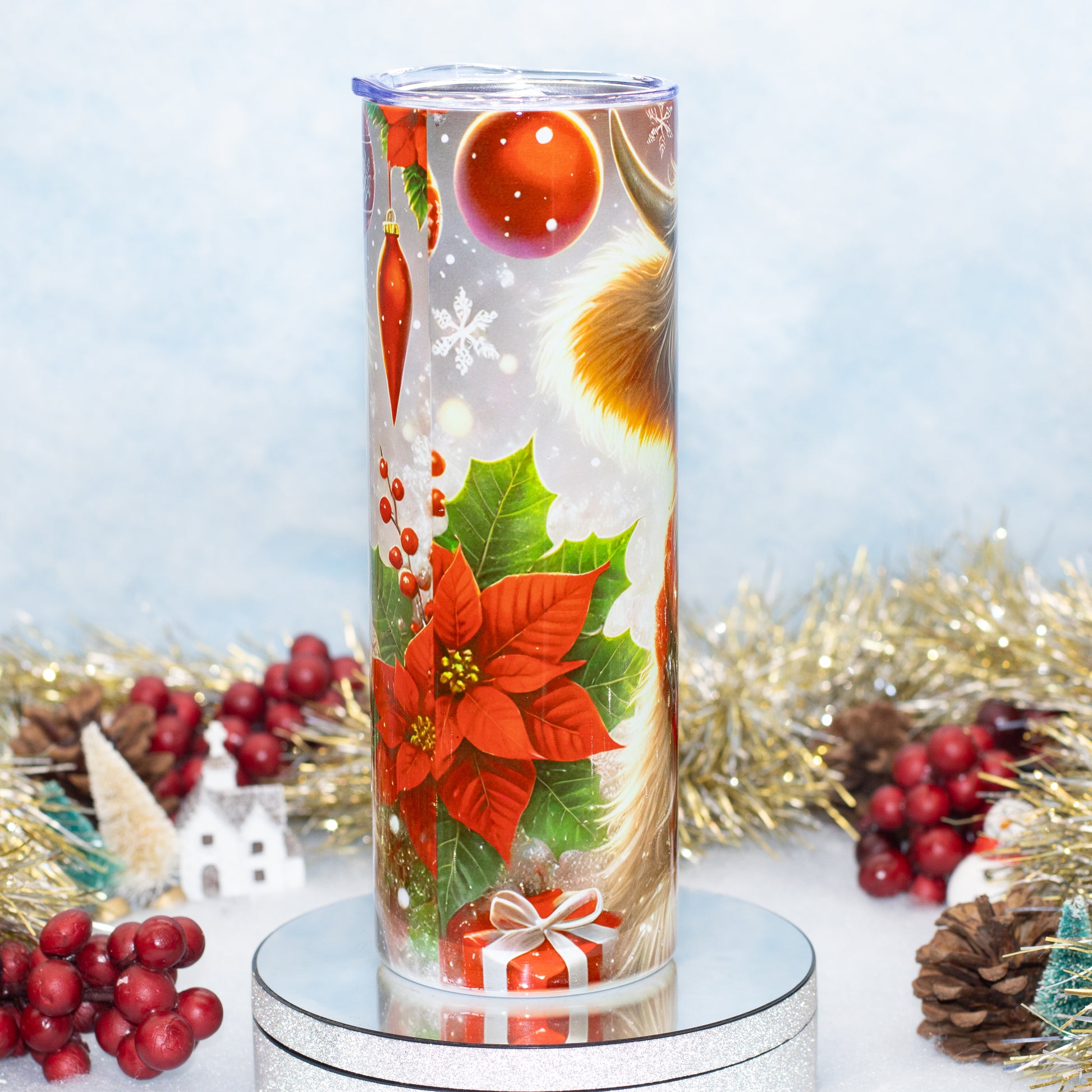A tall cylindrical glass candle with a clear holiday-themed print featuring red poinsettias, green holly leaves, snowflakes, ornaments, and a golden angel wing, displayed on a silver pedestal with festive tinsel and red berries in the background.