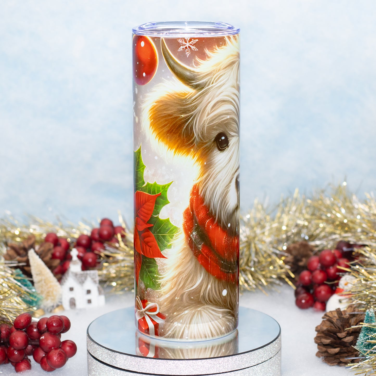 A tall stainless steel tumbler with a printed Christmas design featuring a highland cow in a red sweater, green holly leaves, red berries, and snowflakes, displayed on a silver stand with holiday decorations in the background.