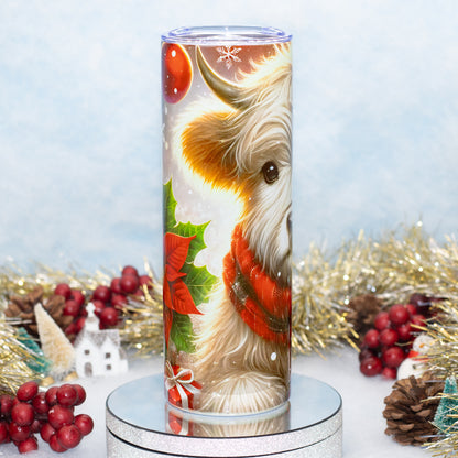 A tall stainless steel tumbler with a printed Christmas design featuring a highland cow in a red sweater, green holly leaves, red berries, and snowflakes, displayed on a silver stand with holiday decorations in the background.