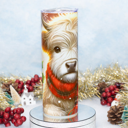 A tall cylindrical tumbler with a clear lid, featuring a printed illustration of a fluffy highland cow wearing a red scarf, displayed on a round silver pedestal with holiday decorations in the background.