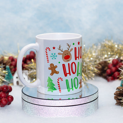 A white ceramic mug with a festive Christmas 'Ho Ho Ho' print featuring a reindeer, candy canes, gingerbread man, and Christmas tree, displayed on a round silver glitter pedestal with holiday decorations in the background.
