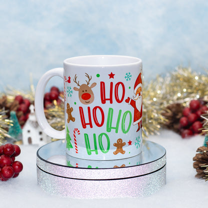 A white ceramic mug with a festive Christmas 'HO HO HO' print, displayed on a round silver glitter pedestal with holiday decorations in the background.