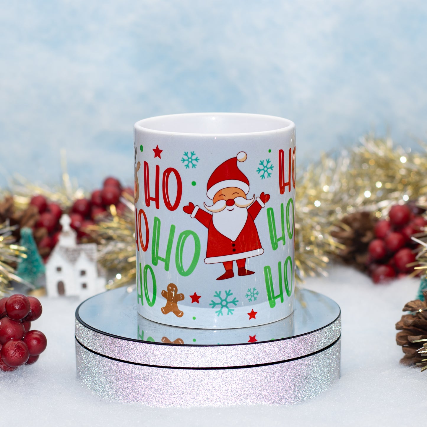 A white ceramic mug with a festive Christmas design featuring Santa Claus, 'HO HO HO' text, snowflakes, and gingerbread cookies, displayed on a round silver glitter pedestal with holiday decorations in the background.
