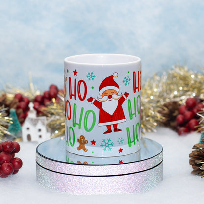 A white ceramic mug with a festive Christmas design featuring Santa Claus, 'HO HO HO' text, snowflakes, and gingerbread cookies, displayed on a round silver glitter pedestal with holiday decorations in the background.
