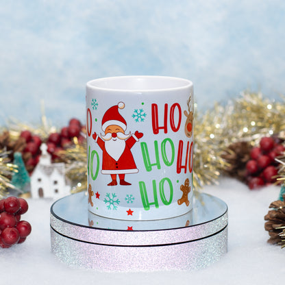 A white ceramic Christmas mug featuring a festive 'Ho Ho Ho' design with a cartoon Santa Claus, snowflakes, and gingerbread men, displayed on a round mirrored pedestal with holiday decorations in the background.