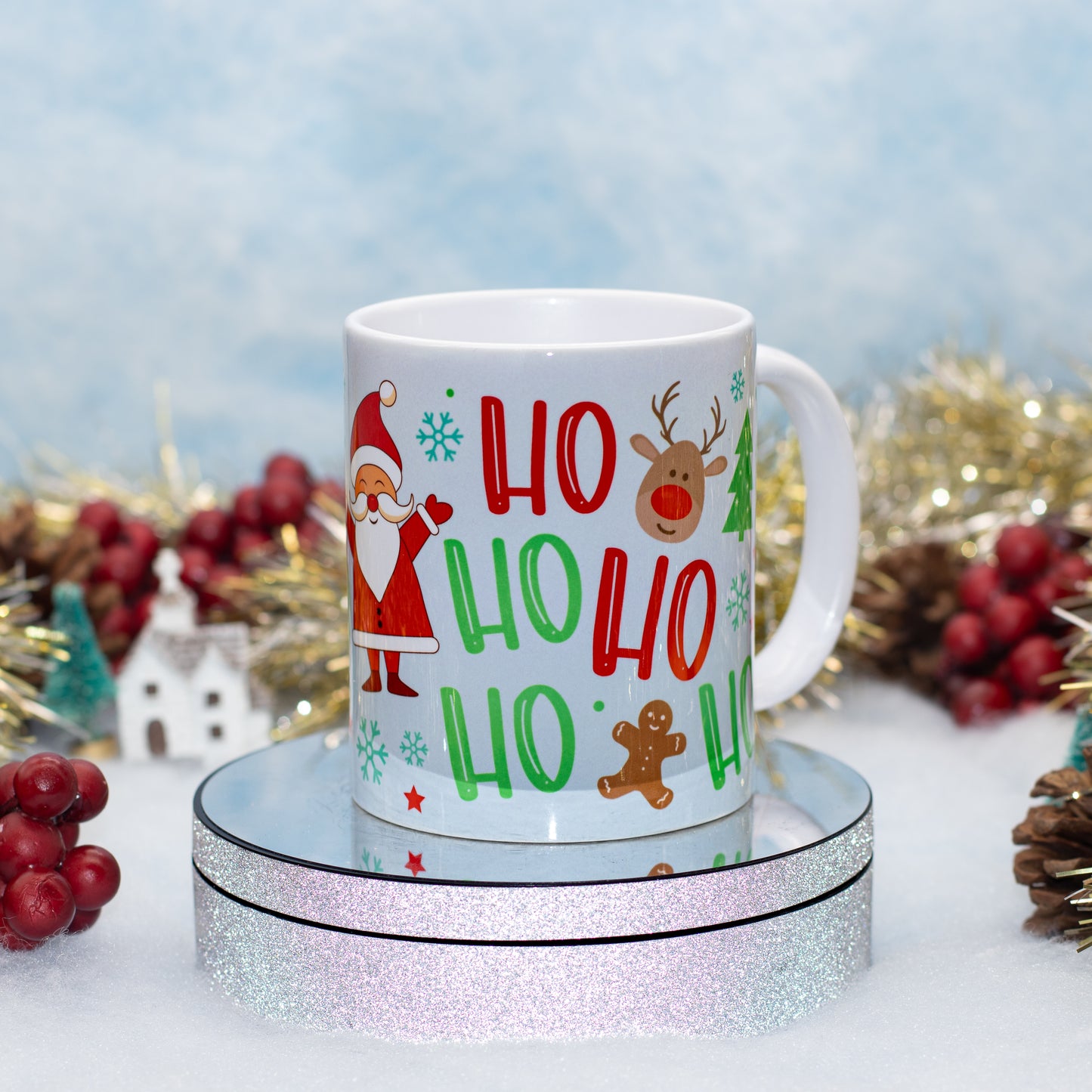 A white ceramic mug with a festive Christmas 'HO HO HO' print featuring Santa Claus, a reindeer, and gingerbread cookies, displayed on a round silver glitter stand with holiday decorations in the background.