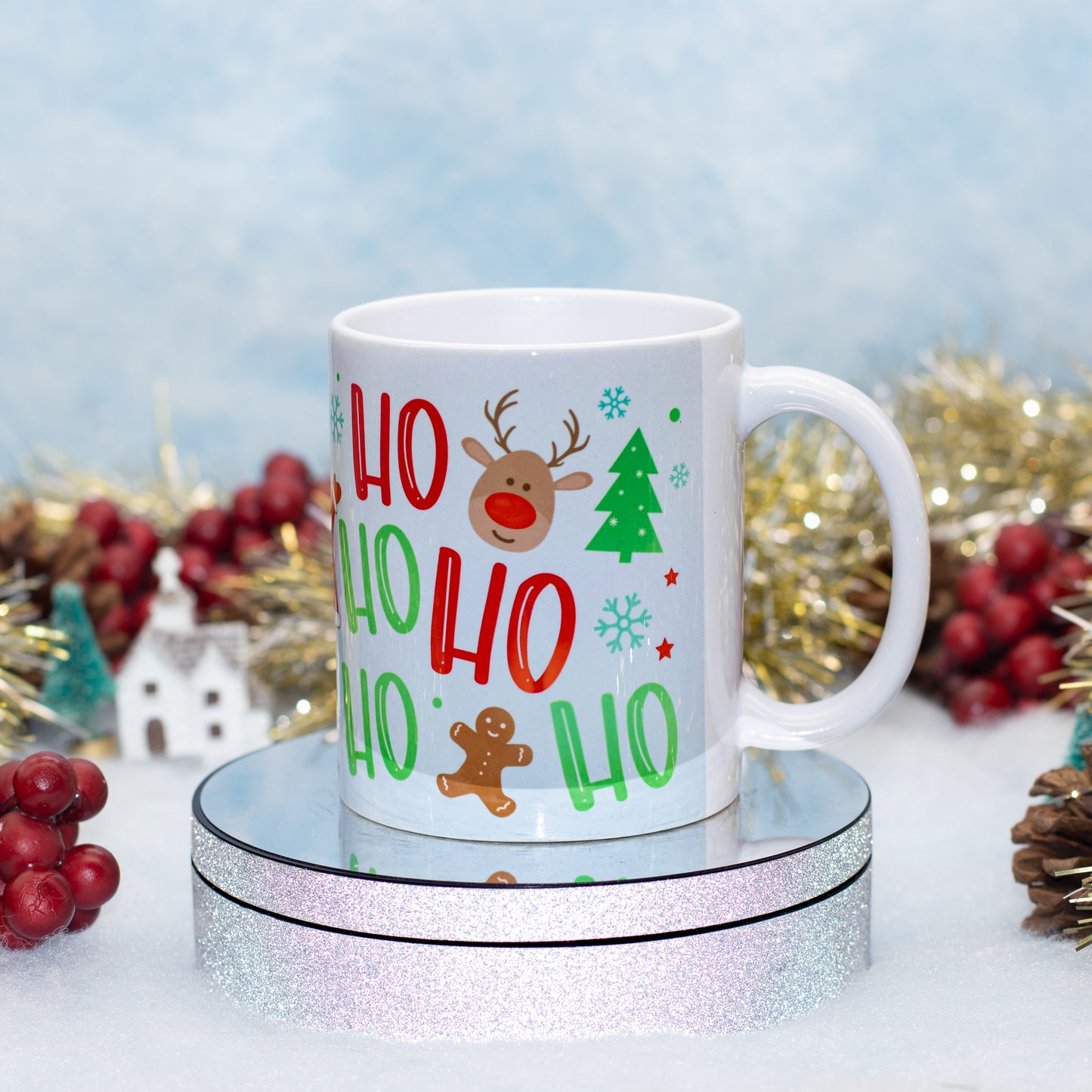 A white ceramic mug with a festive Christmas design featuring the text 'HO HO HO HO HO HO HO' in red and green, a brown reindeer, a green Christmas tree, and a gingerbread man, displayed on a round mirrored pedestal with holiday decorations in the background.