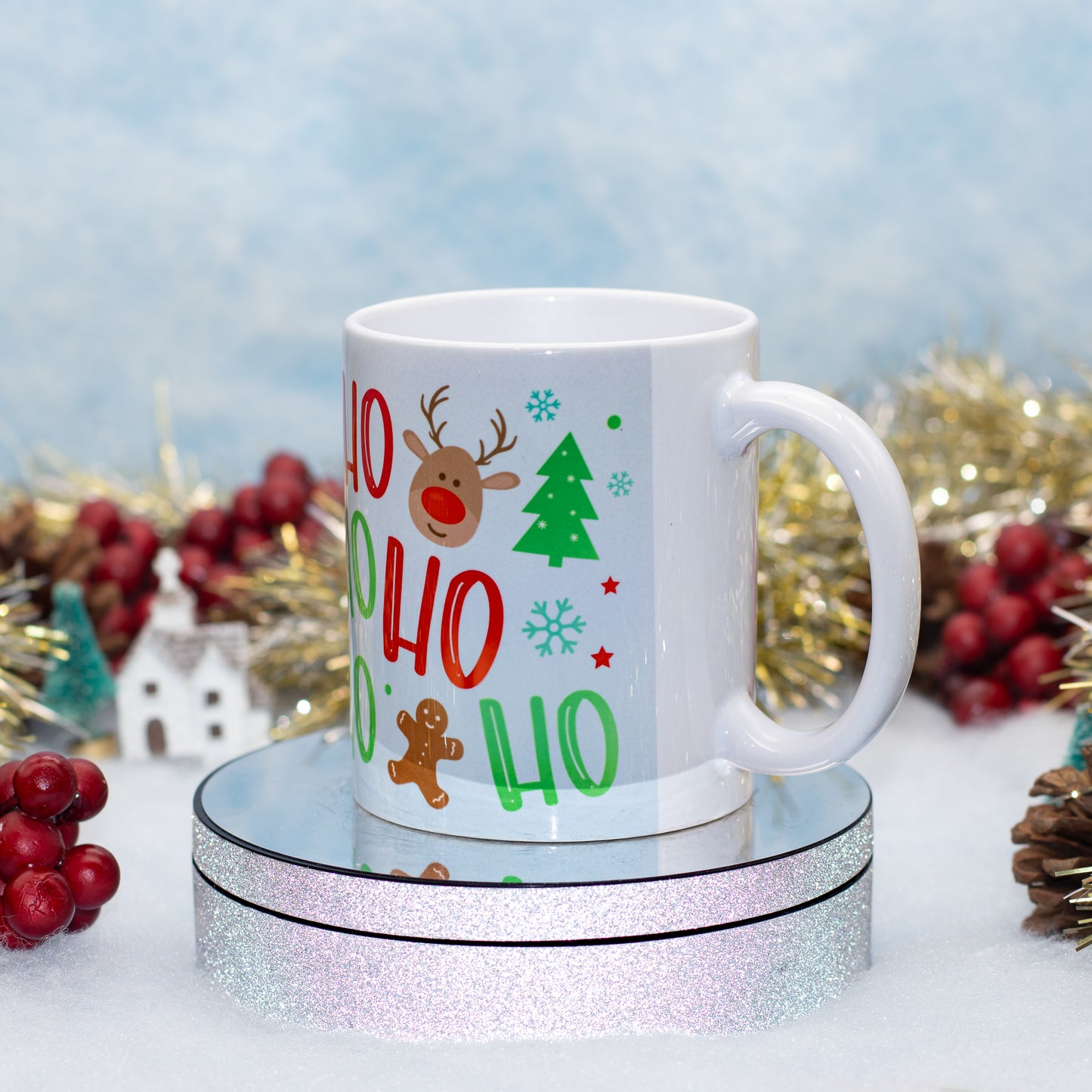 A white ceramic mug with a festive 'Ho Ho Ho' holiday design, featuring a reindeer, Christmas tree, and gingerbread man, displayed on a round glittery stand against a snowy backdrop with holiday decorations.