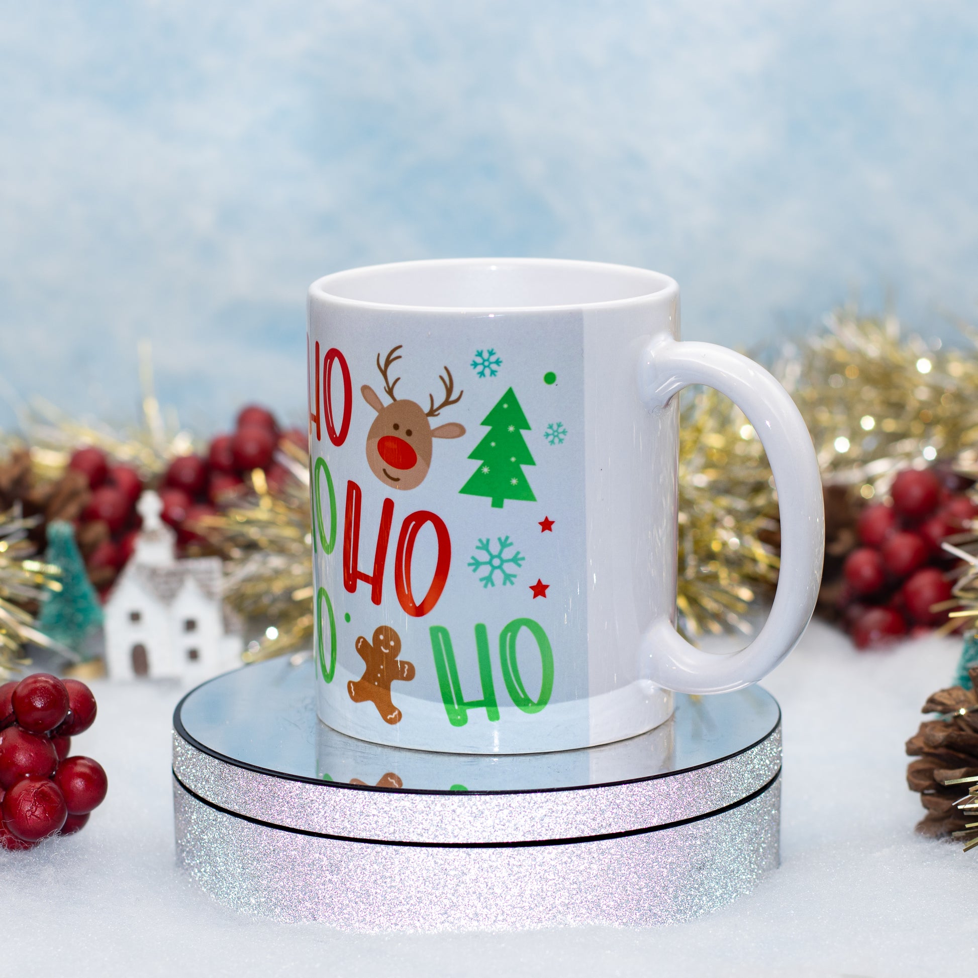 A white ceramic mug with a festive 'Ho Ho Ho' holiday design, featuring a reindeer, Christmas tree, and gingerbread man, displayed on a round glittery stand against a snowy backdrop with holiday decorations.