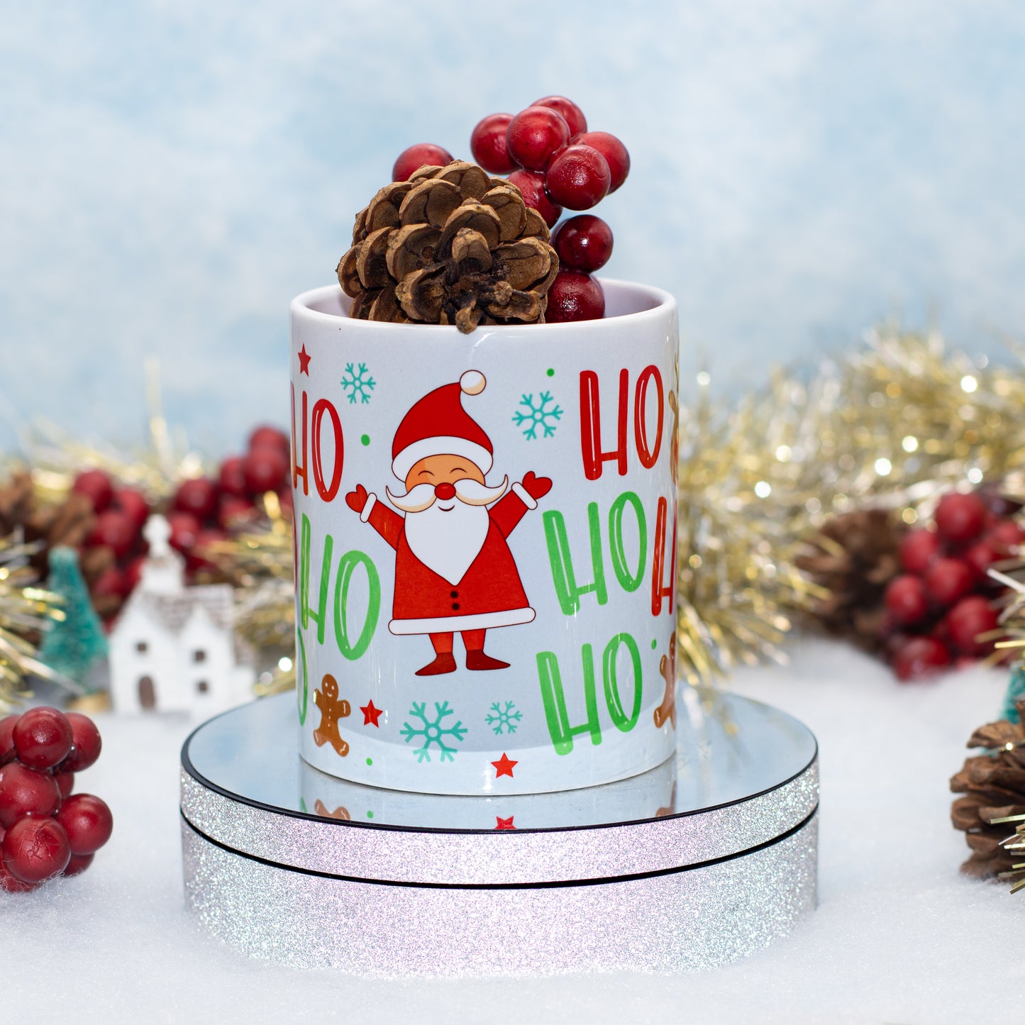 A white ceramic mug with a festive 'Ho Ho Ho' Santa Claus design, styled on a round mirrored pedestal with pinecones and red berries against a soft blue holiday backdrop.